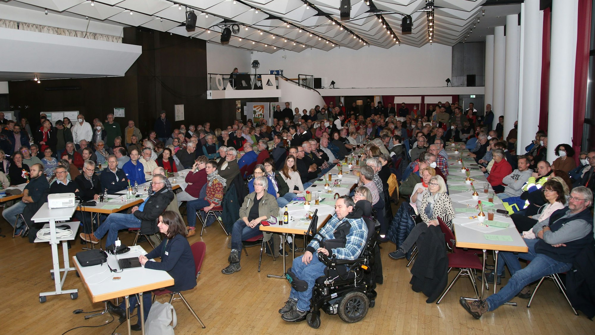 In einer Halle sitzen zahlreiche Menschen an langen Tischen. Einige haben keinen Platz gefunden und stehen am Rand.