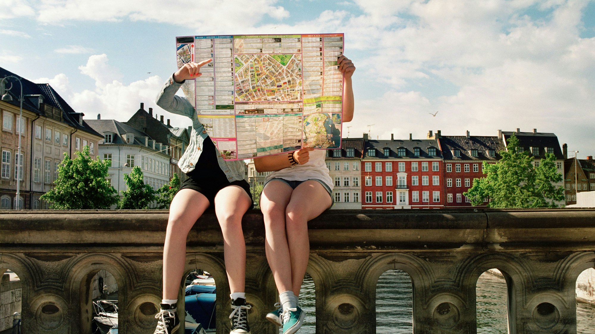 Zwei Teenager sitzen auf einer Brücke und halten eine Landkarte vor ihre Gesichter.
