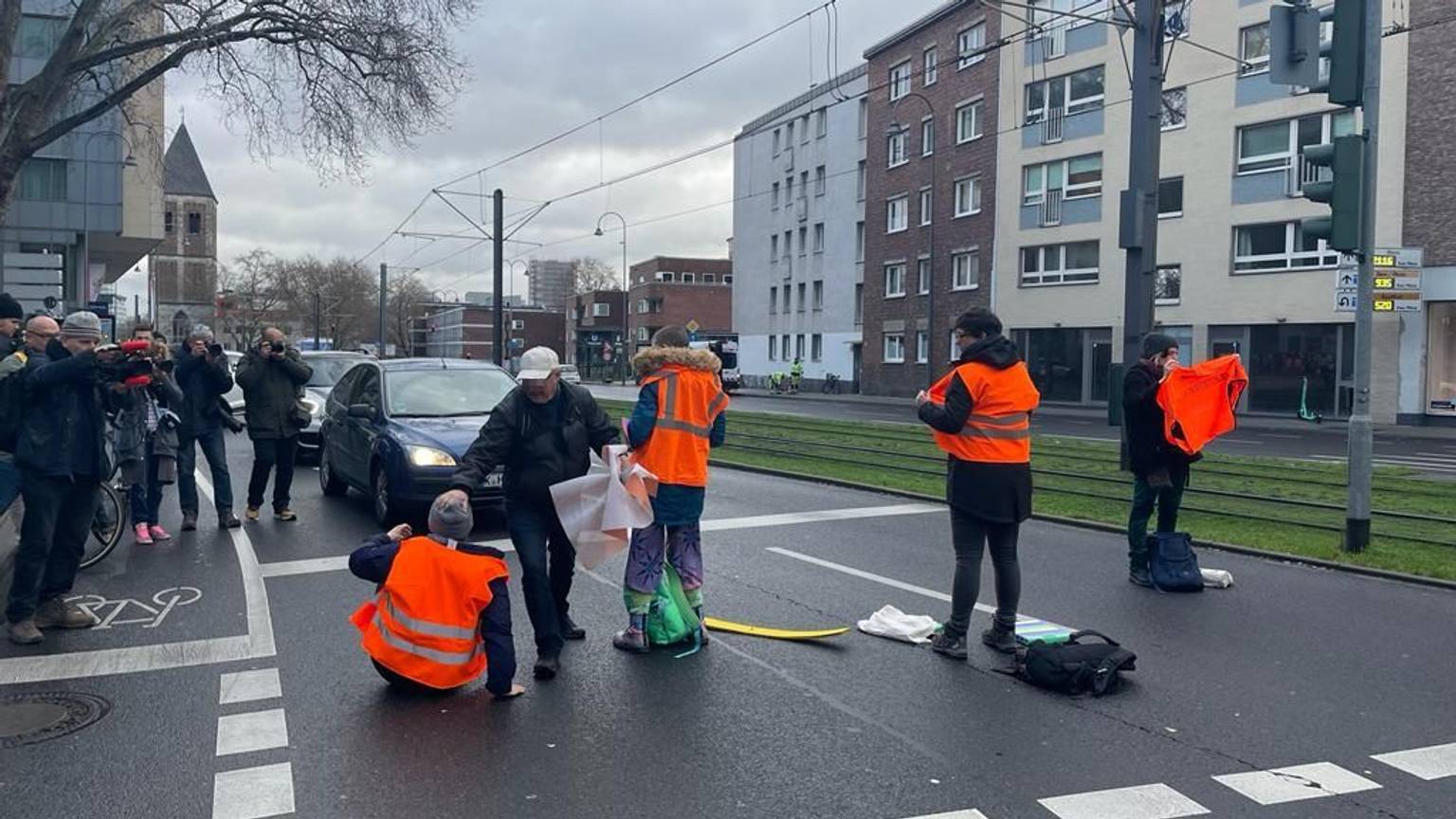 Ein Mann mit grauer Jacke greift Klimaaktivisten mit orangen Warnwesten bei einer Straßensperre in der Kölner Innenstadt an.