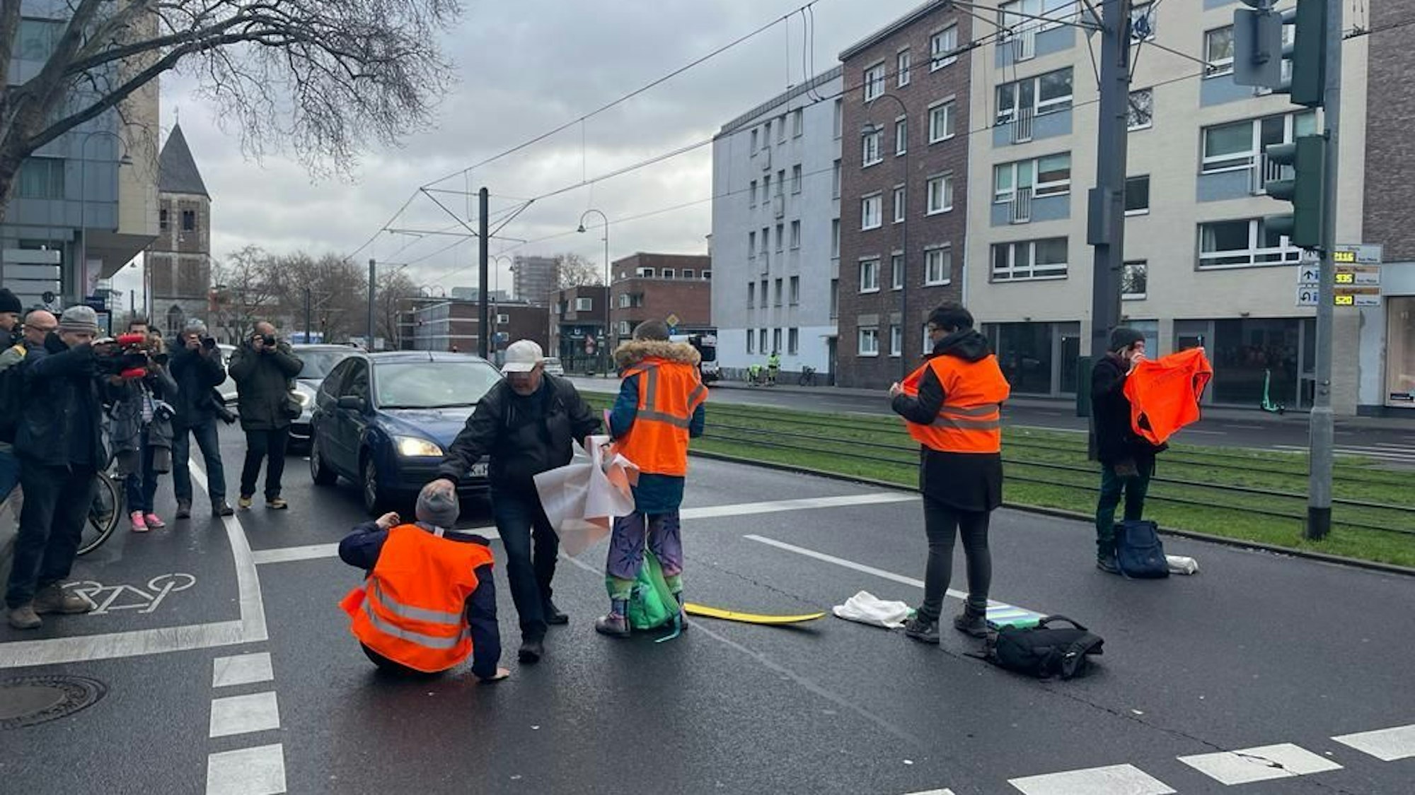 Ein Mann mit grauer Jacke greift Klimaaktivisten mit orangen Warnwesten bei einer Straßensperre in der Kölner Innenstadt an.