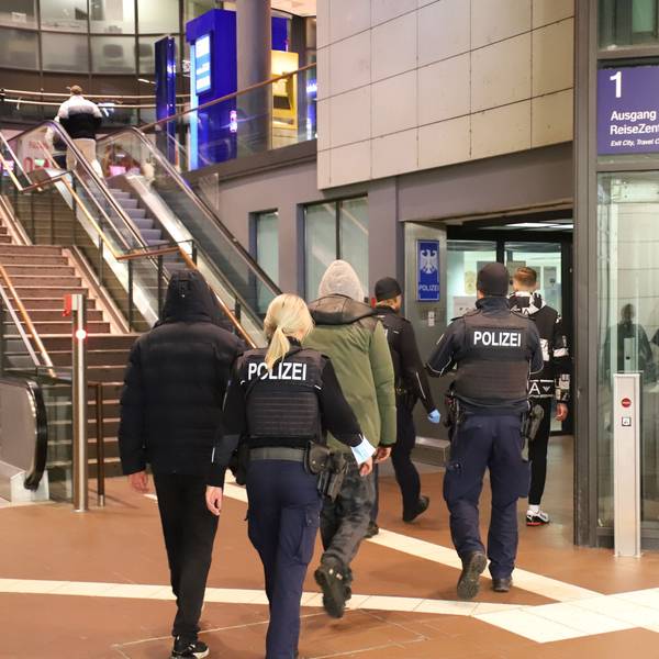 Im Siegburger Bahnhof vor den Treppen und Rolltreppen und neben dem Aufzug gehen zwei uniformierte Beamte. Sie sind von hinten zu sehen, eine blonde Frau mit Pferdeschwanz und ein Beamter mit Mütze begleiten drei Männer, die Kapuzenjacken tragen.
