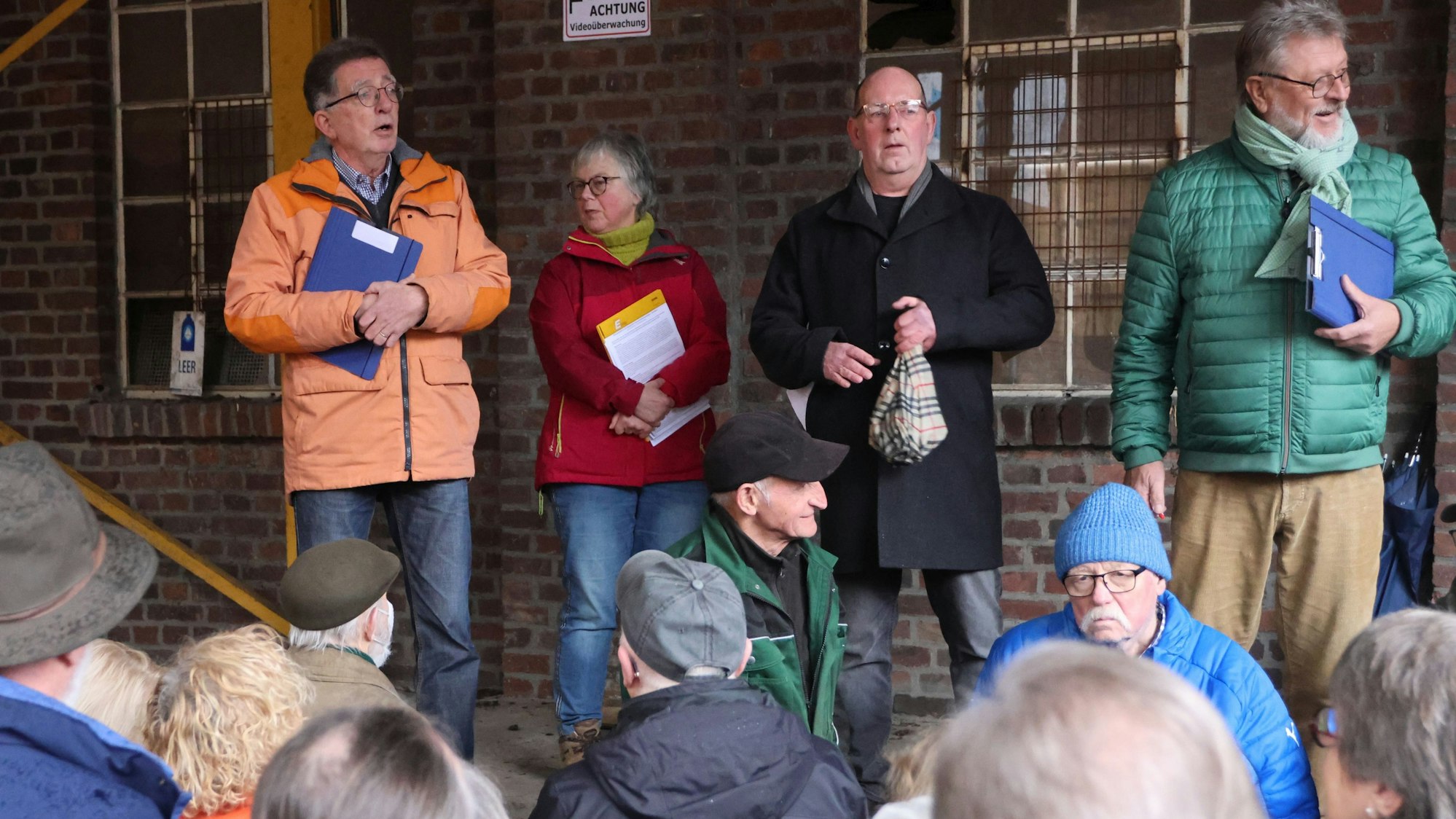Drei Männer und eine Frau stehen mit Ordnern in der Hand vor einer Reihe Menschen.