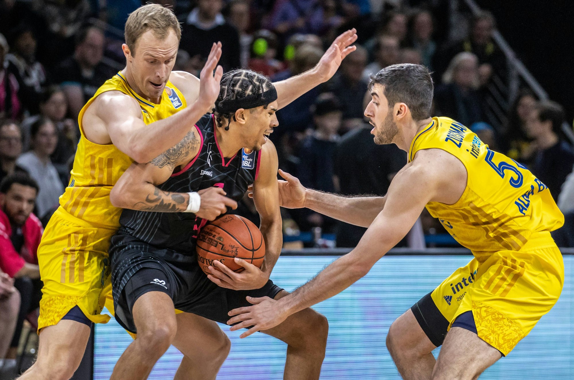 Zwei Basketballer in gelben Trainingsanzügen versuchen einem gegenerischen Spieler den Ball abzunehmen.