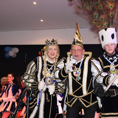 Ein Dreigestirn steht, gekleidet in den Ornaten, auf der Bühne. Der Prinz in der Mitte spricht in ein Mikrofon.