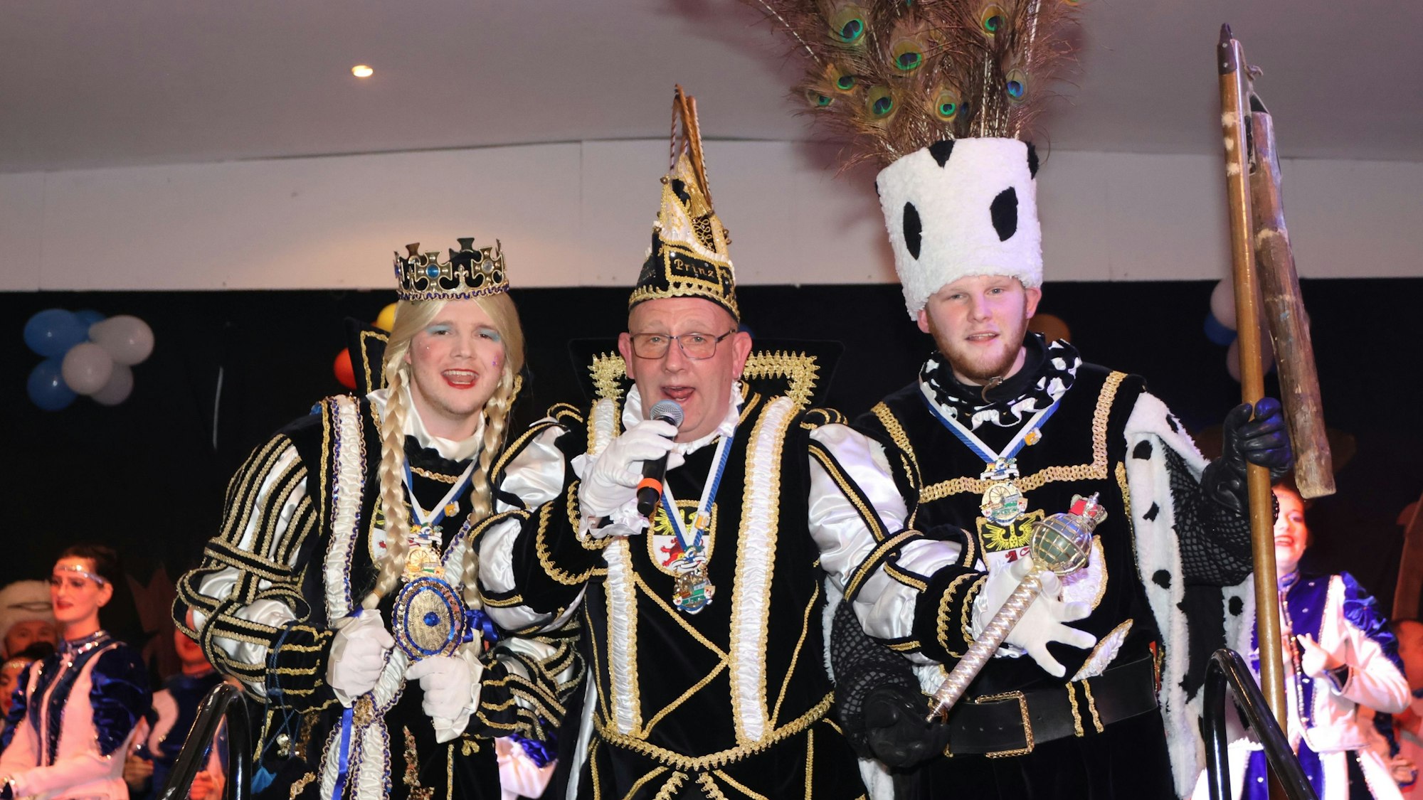 Ein Dreigestirn steht, gekleidet in den Ornaten, auf der Bühne. Der Prinz in der Mitte spricht in ein Mikrofon.