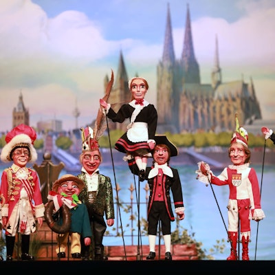 Verschiedene Puppen stehen vor einer Leinwand, die den Kölner Dom abbildet.