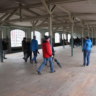 Einige Menschen laufen durch eine leere, nicht mehr genutzte Fabrikhalle mit großen Fenstern.