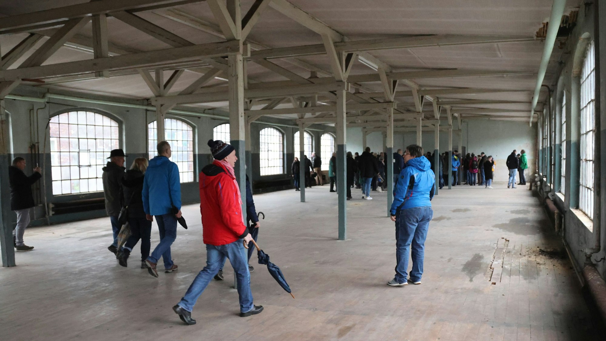 Einige Menschen laufen durch eine leere, nicht mehr genutzte Fabrikhalle mit großen Fenstern.