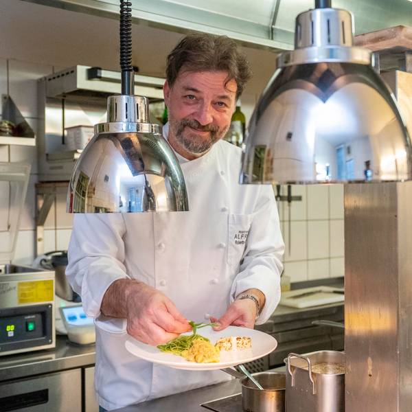 Roberto Carturan steht in Kochkleidung in seiner Küche und hält einen Teller mit Essen in der Hand
