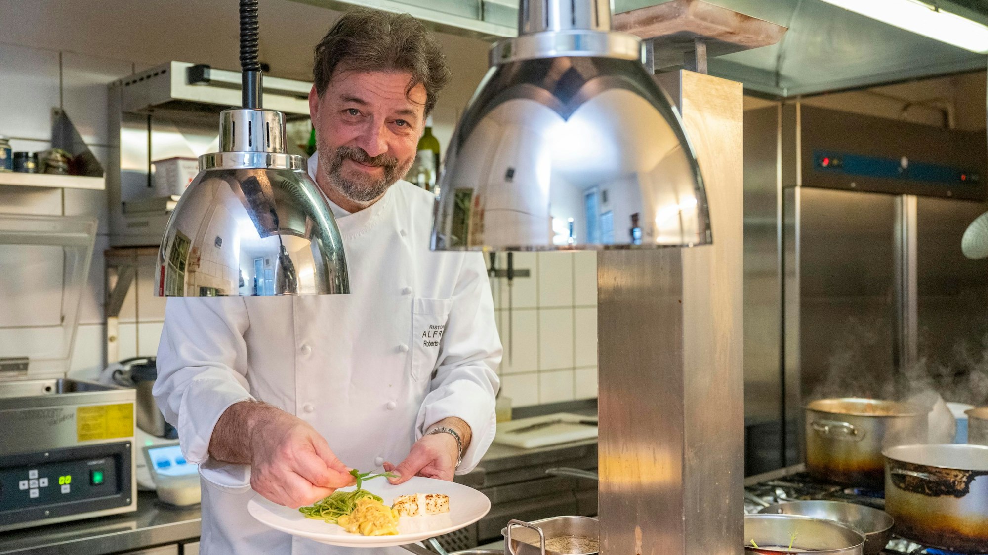 Roberto Carturan steht in  Kochkleidung in seiner Küche und hält einen Teller mit Essen in der Hand