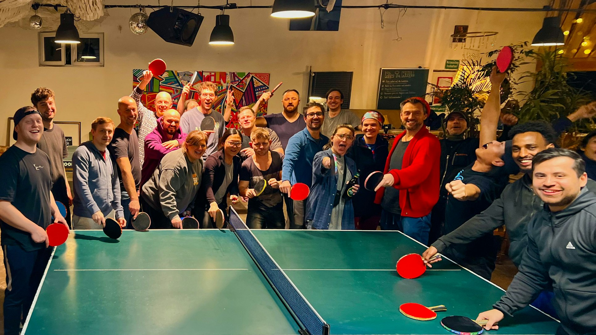 Etwa 20 junge Leute umringen um eine Tischtennissplatte, viele von ihnen in Jubelpose.