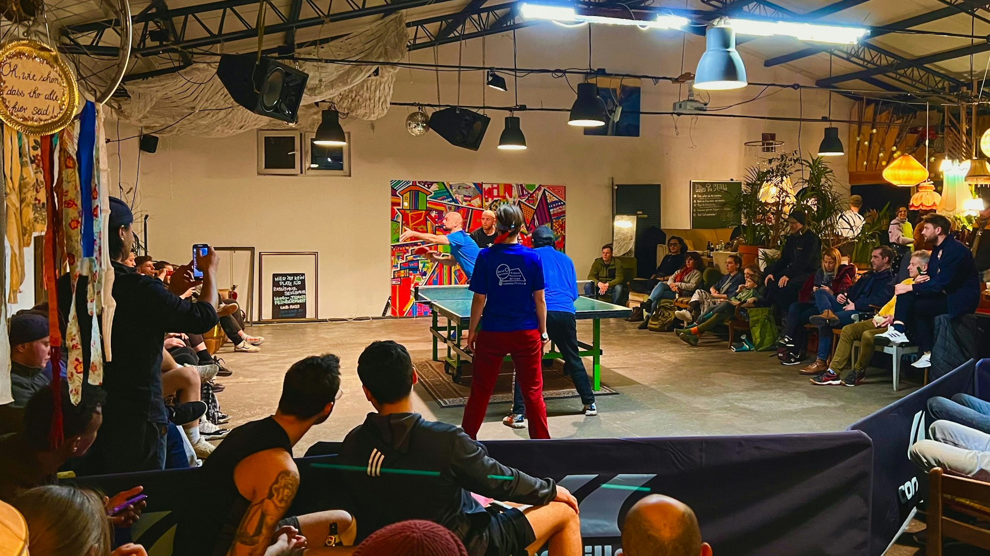In einer großen Halle mit Sofas und vielen Lampen, die von der Decke hängen, sitzen Zuschauer um eine Tischtennisplatte herum, an der gerade ein Doppel stattfindet.