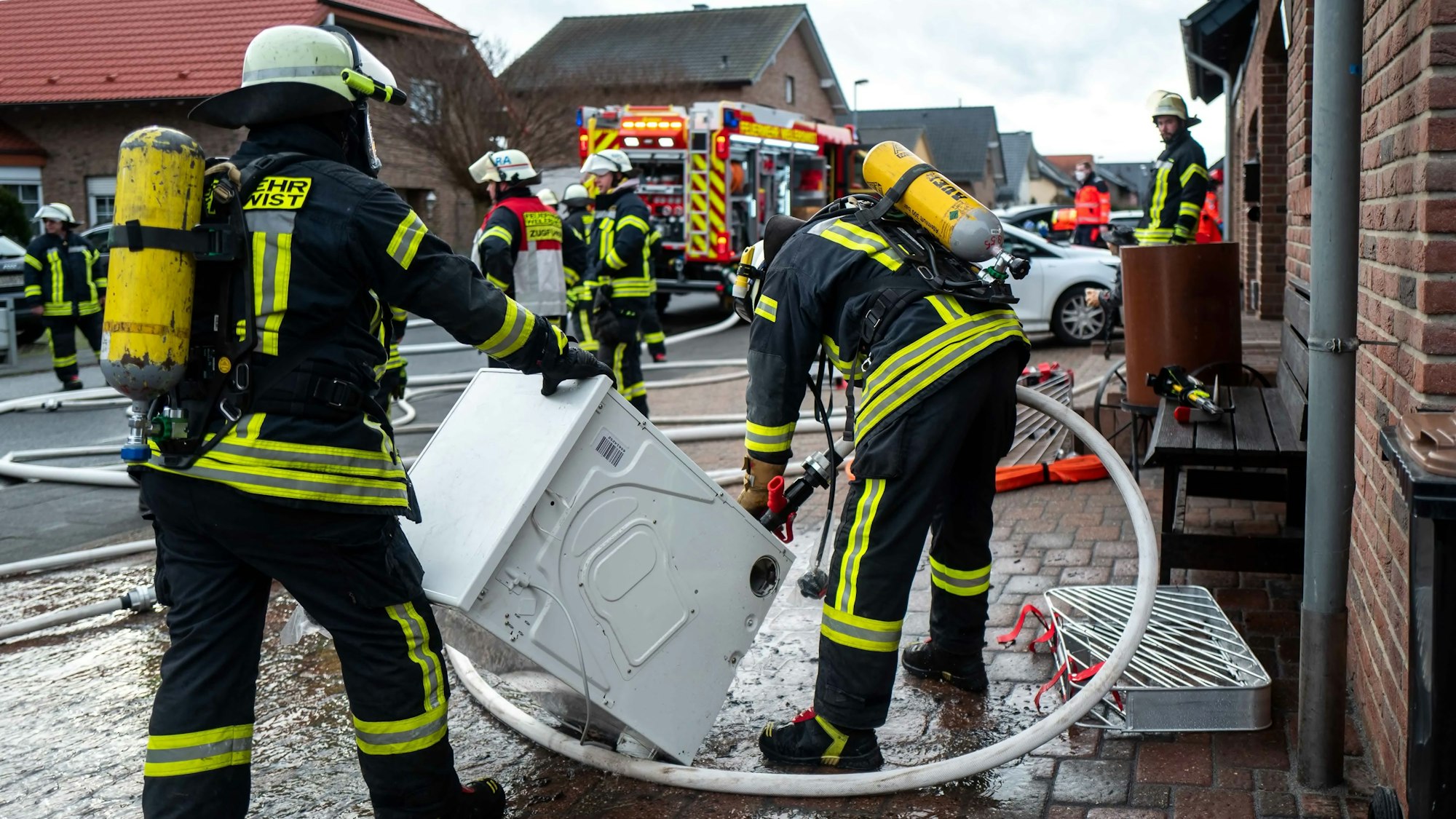 Zwei Feuerwehrleute löschen vor dem Haus einen Trockner. Im Hintergrund sind weitere Einsatzkräfte und ein Feuerwehrauto zu sehen.