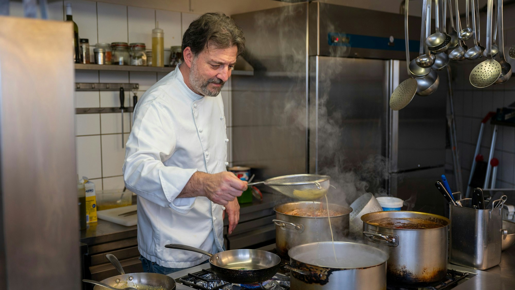 Mann in weißer Kochjacke steht vor einem Herd mit vielen Töpfen
