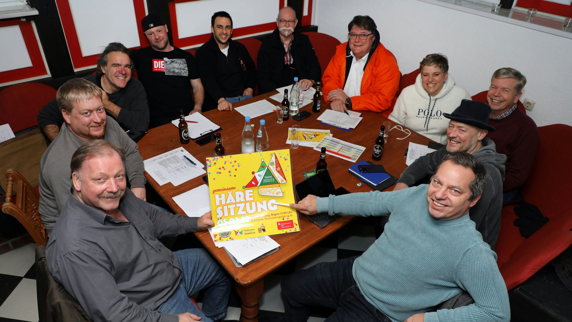Eine Gruppe Männer sitzt am Tisch an einer Kneipe, zwei halten das Plakat für die neue Bergisch Gladbacher Karnevalssitzung in die Kamera.