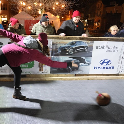 Zu sehen ist die Kunststoffbahn, die beim Bergneustädter Wintermärchen die Eisbahn ersetzt. Im Vordergrund wirft eine Frau mit dicker Jacke, Schal und Mütze einen Eisstock.
