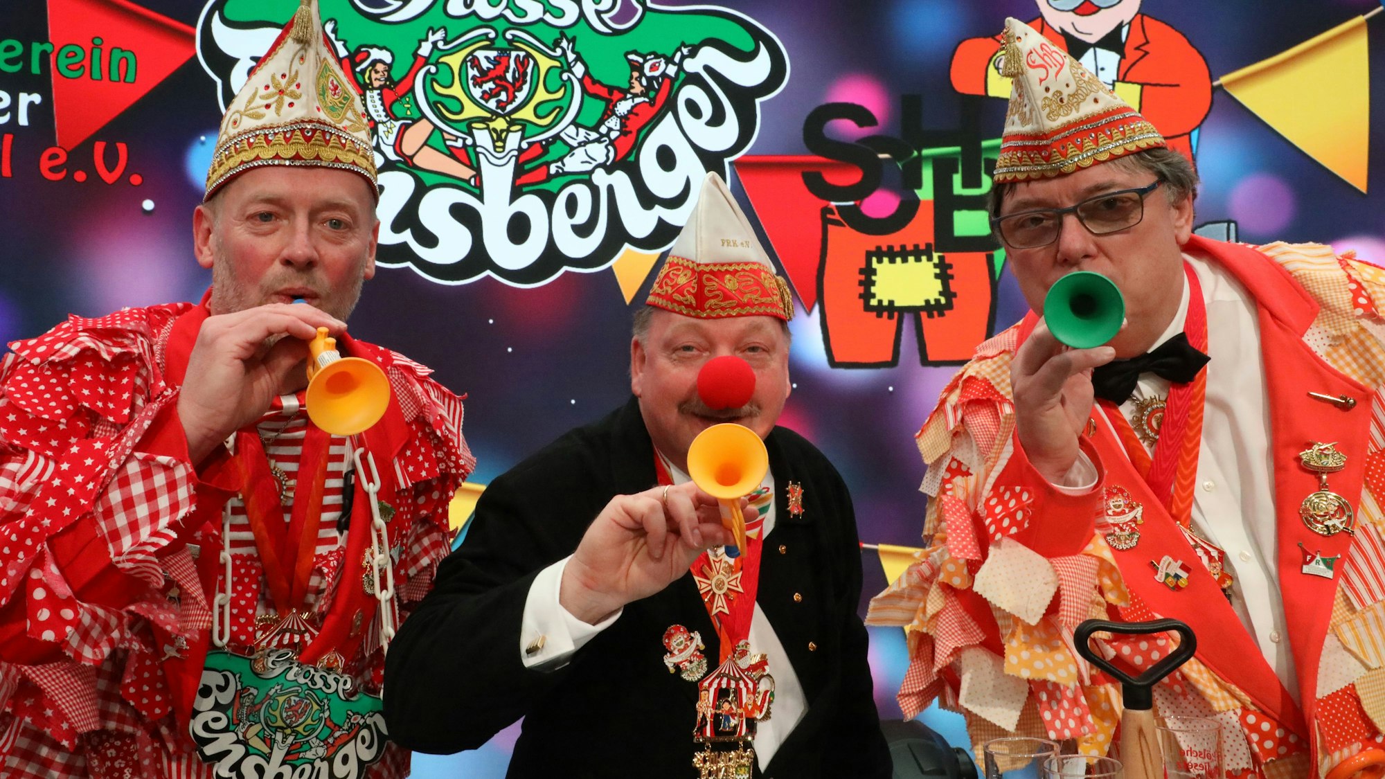 Rainer Furth-Quernheim (Große Bensberger), Harald Hahn (Förderverein Refrather Karneval) und Schlader-Botze-Chef Michael „Mio“ Oppermann (v.l.) stehen in Karnevalsklamotten mit bunten Tröten vor buntem Hintergrund.