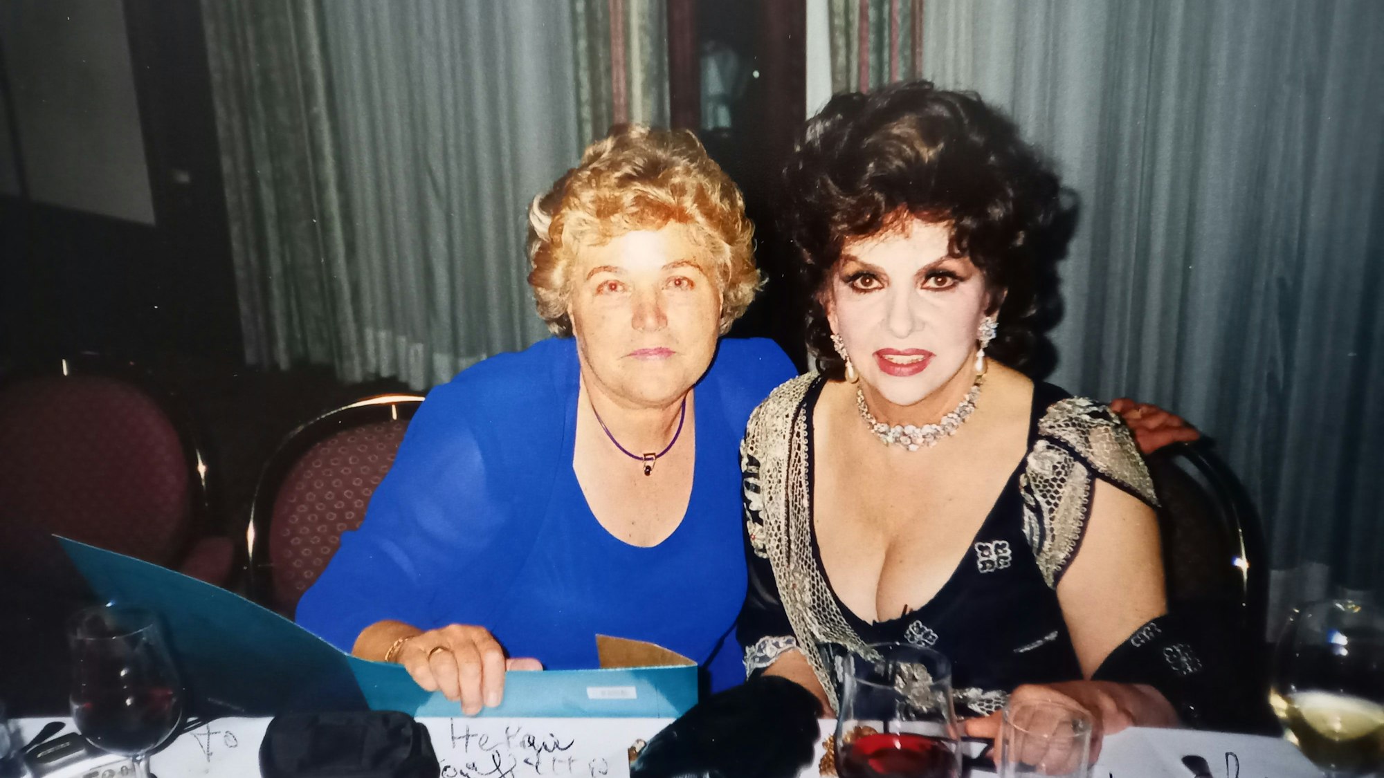Helga Schlösser hat den Arm um Gina Lollobrigida gelegt. Die Frauen sitzen an einem Tisch mit Weingläsern.