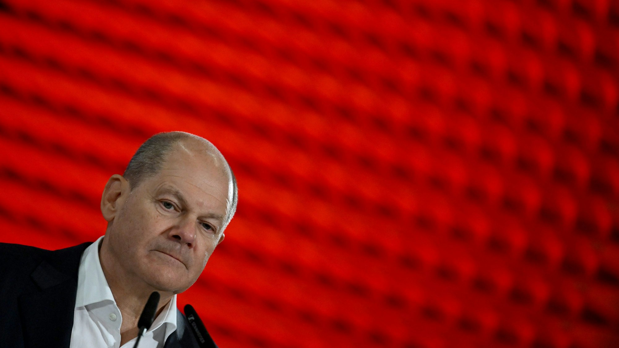 Scholz ist schräg unten links im Bild zu sehen, nachdenkliche Miene. Der Hintergrund ist rot.