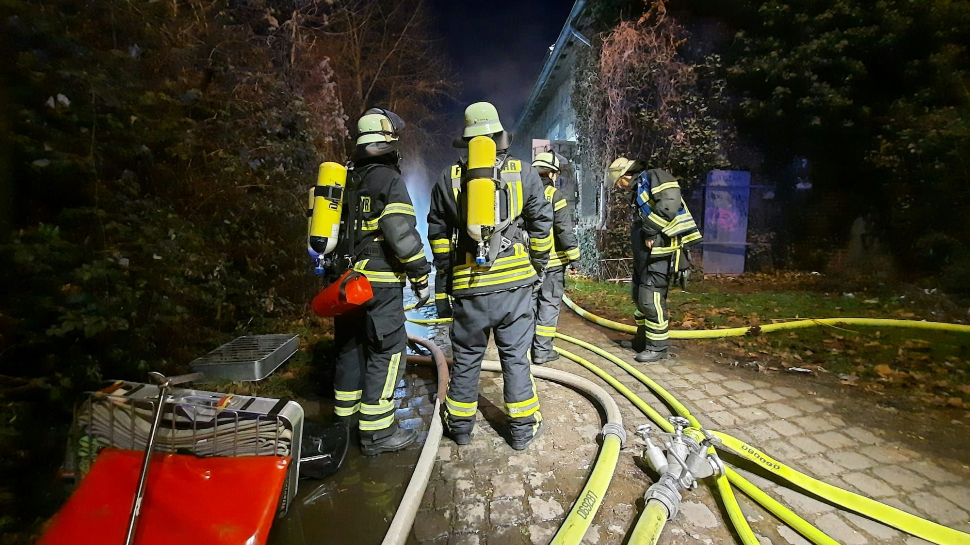 Eine Gruppe von Feuerwehrleuten mit schwerem Atemschutz stehen vor dem Gebäude; auf dem Boden liegen mehrere gelbe Löschschläuche.