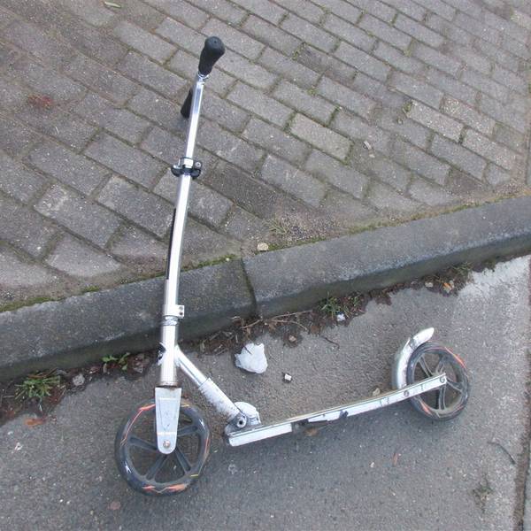 Ein leicht verbogener Roller liegt am Bordstein eines Gehwegs neben einer Straße.
