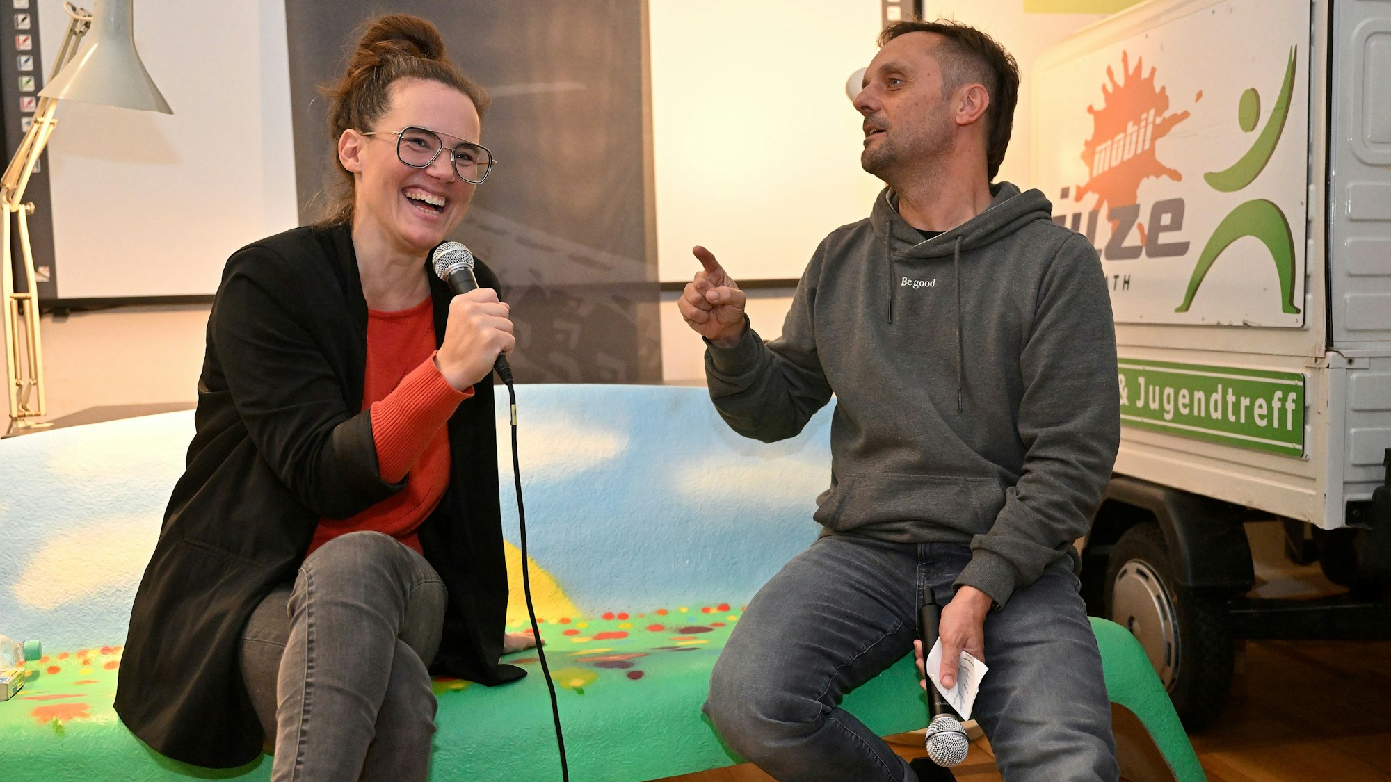 Katja Diehl sitzt neben Moderator Johannes Thies und lacht.