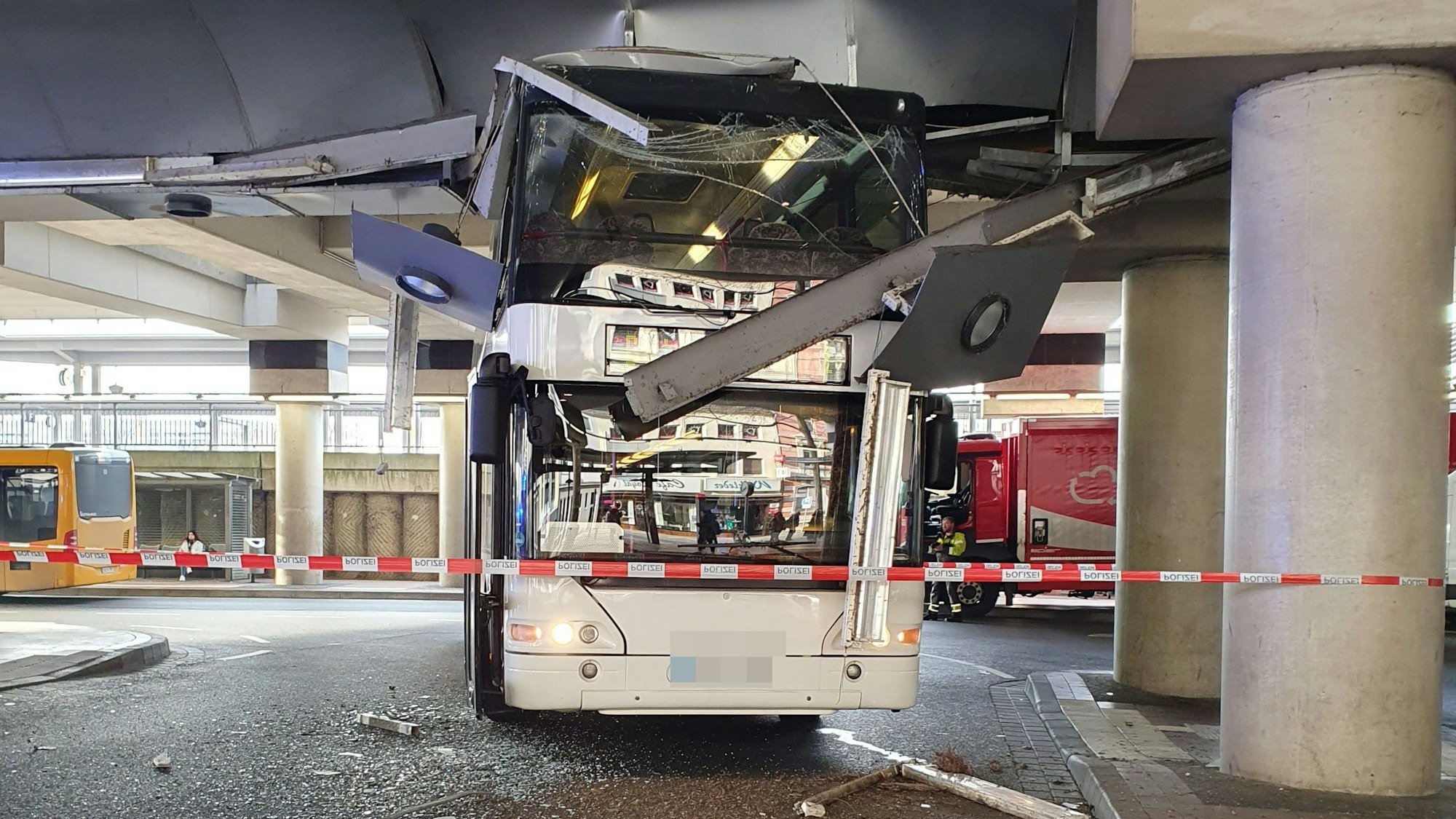 Ein Doppeldeckerbus hat eine zerstörte Scheibe, Teile der Decke hängen runter, der Bereich ist mit einem Polizeiband abgesperrt.