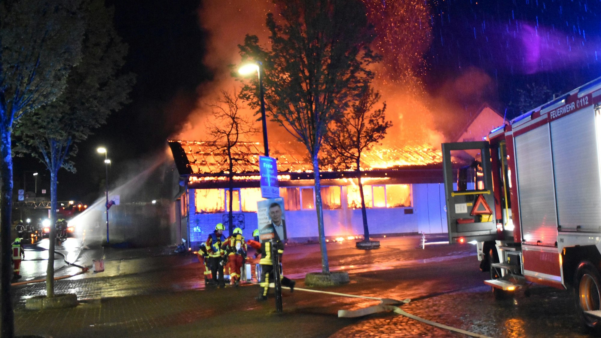 Eine Markthalle steht nachts lichterloh in Flammen. Feuerwehrleute versuchen mit Wasser, die Flammen zu löschen.