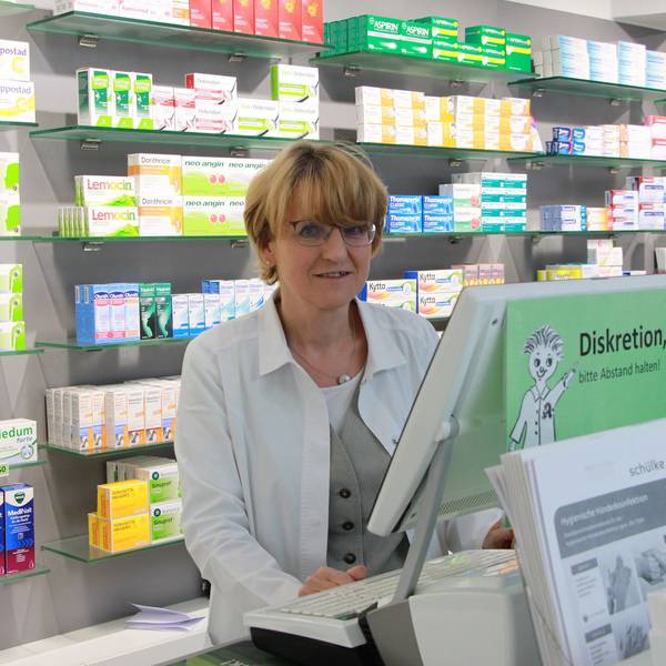 Eine Frau mit blonden Haaren und Brille im weißen Kittel steht in einer Apotheke.