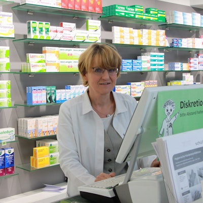 Eine Frau mit blonden Haaren und Brille im weißen Kittel steht in einer Apotheke.
