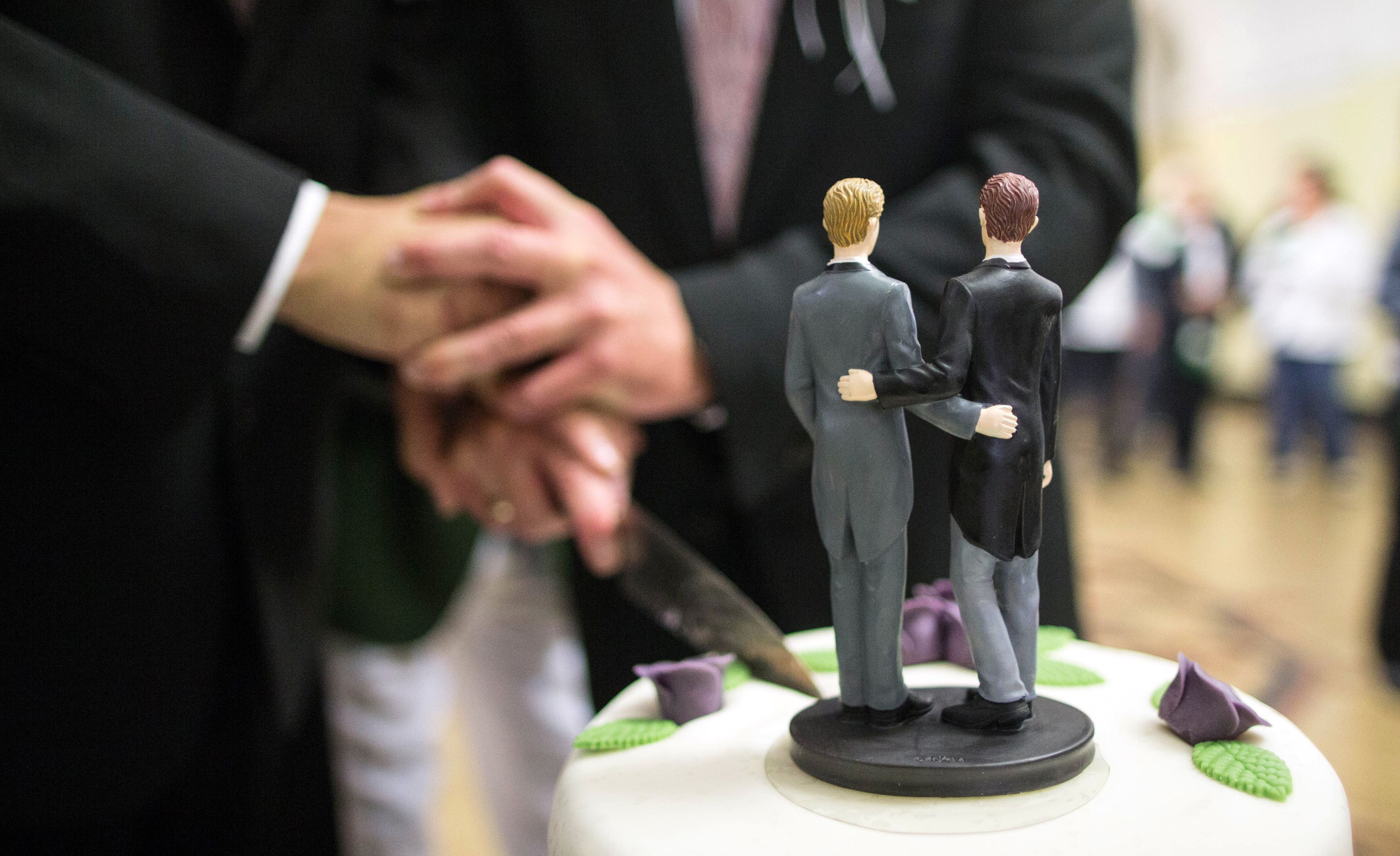 Zwei Männer schneiden eine Hochzeitstorte an, auf der Figren von ihnen sich die Arme umlegen.
