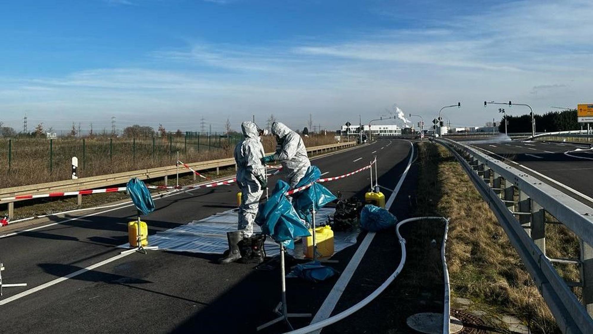 Feuerwehrleute kümmern sich um die ausgetreten Flüssigkeit auf der B 265n in Hürth. Sie tragen weiße Schutzanzüge.