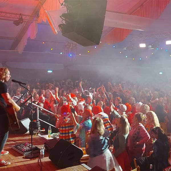 Eine Band steht auf der Bühne, die Fans in Karnevalskostümen feiern in einem Festzelt.