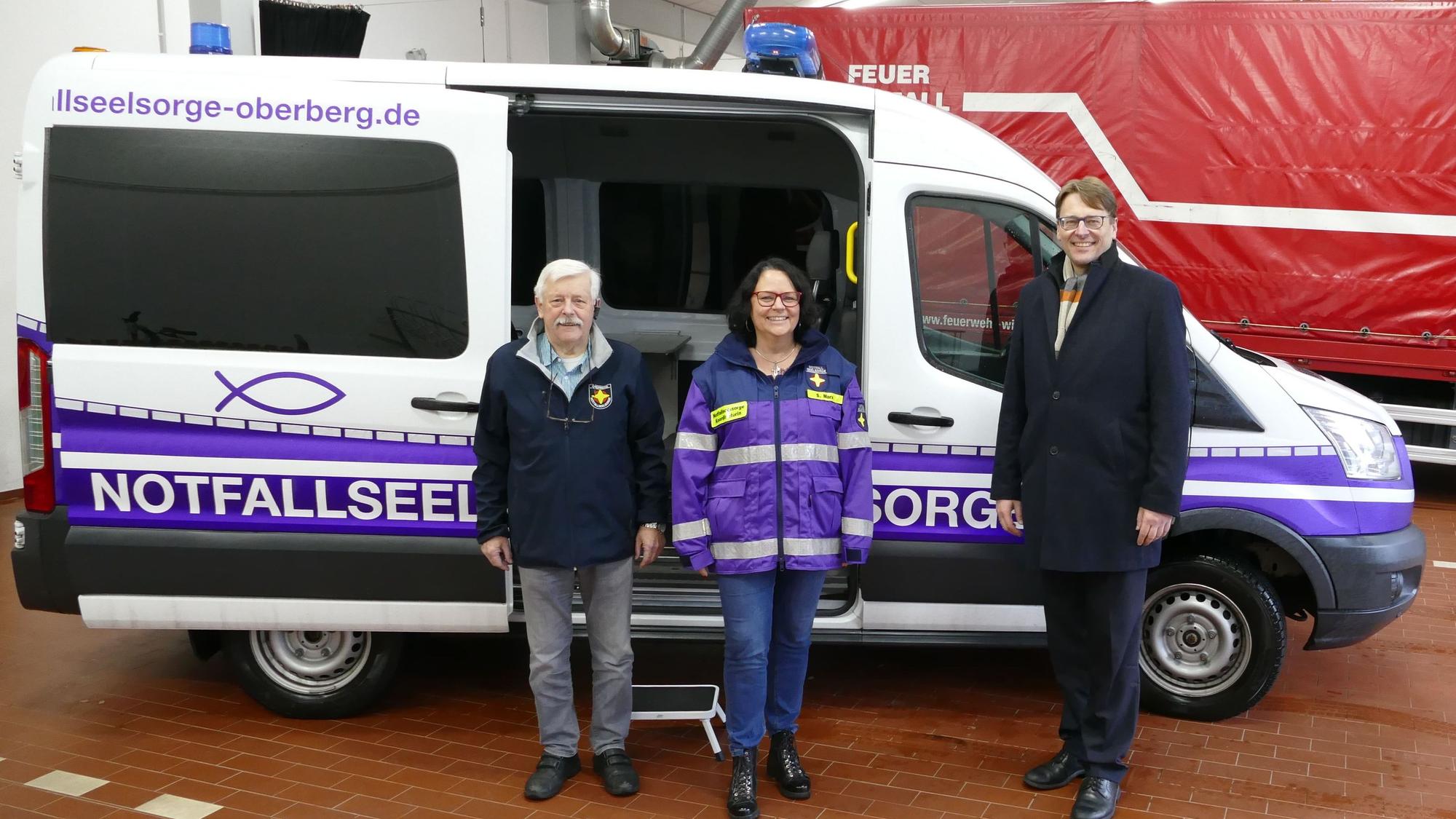 Zwei Männer und eine Frau stehen vor einem weißen Kleinbus der Notfallseelsorge mit Blaulicht.