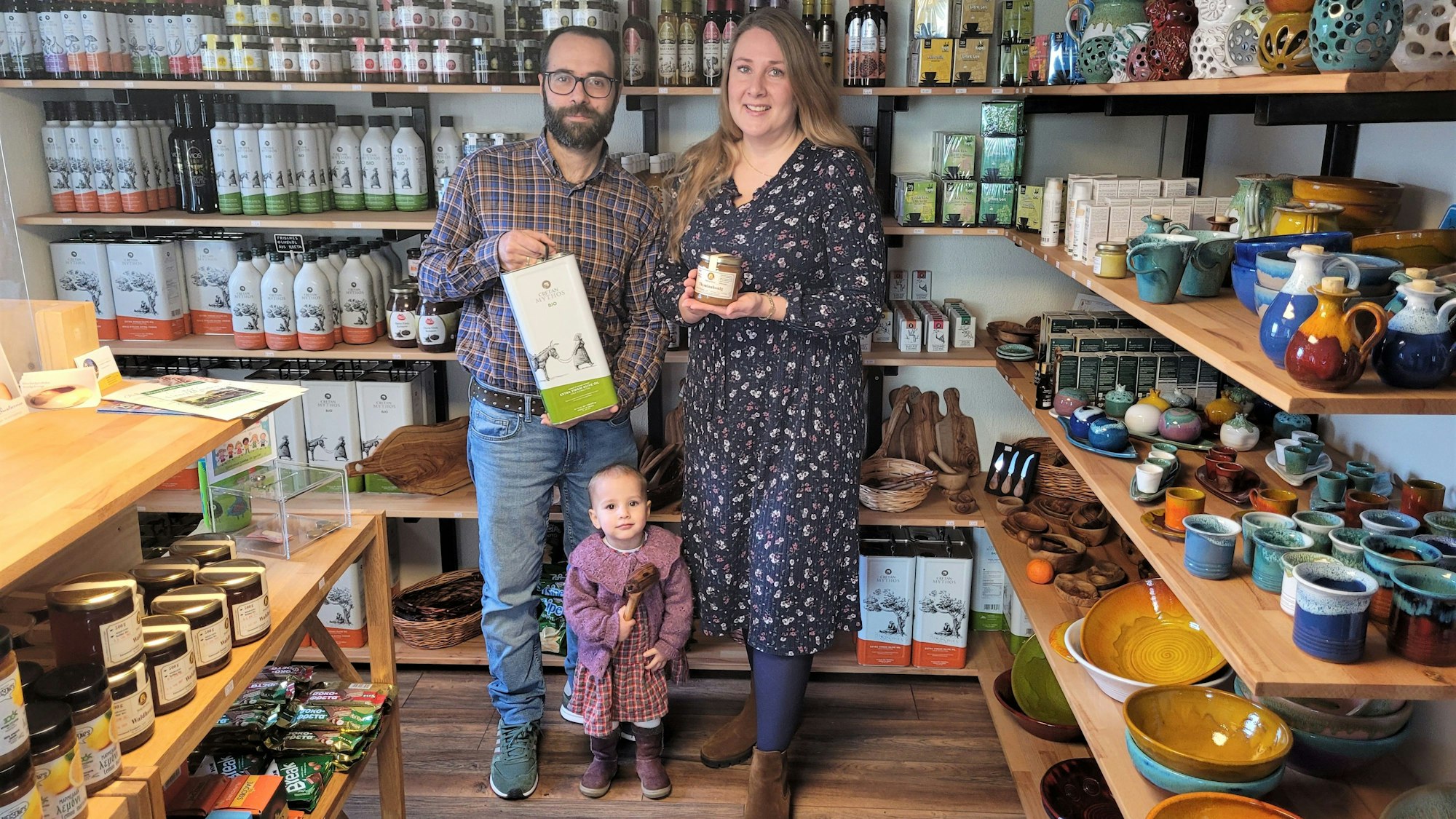 Ein Mann und eine Frau stehen mit ihrer kleinen Tochter in ihrem Geschäft mit griechischen Spezialitäten.