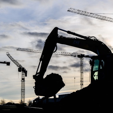 Ein Bagger und einige Baukräne im Hintergrund vor einem bewölkten Himmel