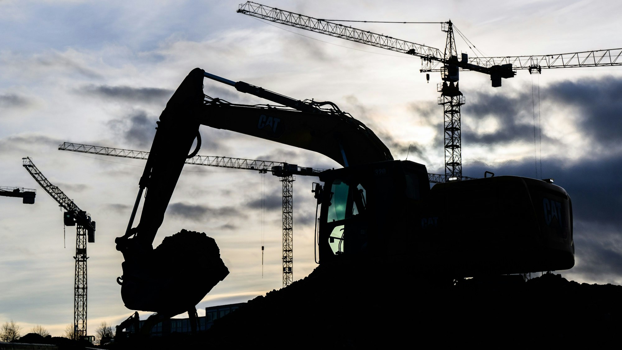 Ein Bagger und einige Baukräne im Hintergrund vor einem bewölkten Himmel