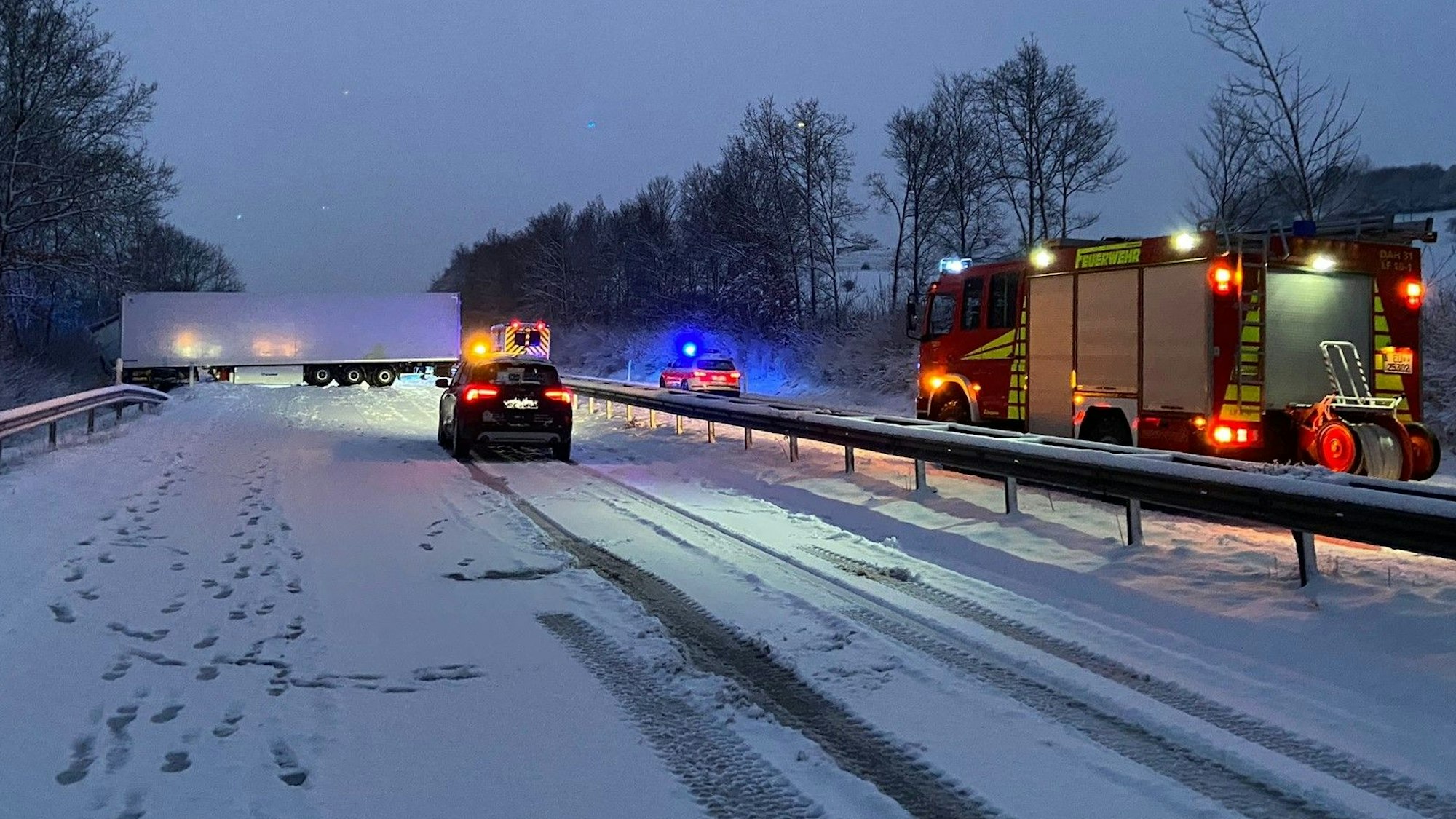 Ein Lastzug hat sich auf der verschneiten B51 quergestellt und blockiert die Fahrbahn. Ein Feuerwehrfahrzeug sichert in der einsetzenden Dämmerung den Bereich ab.