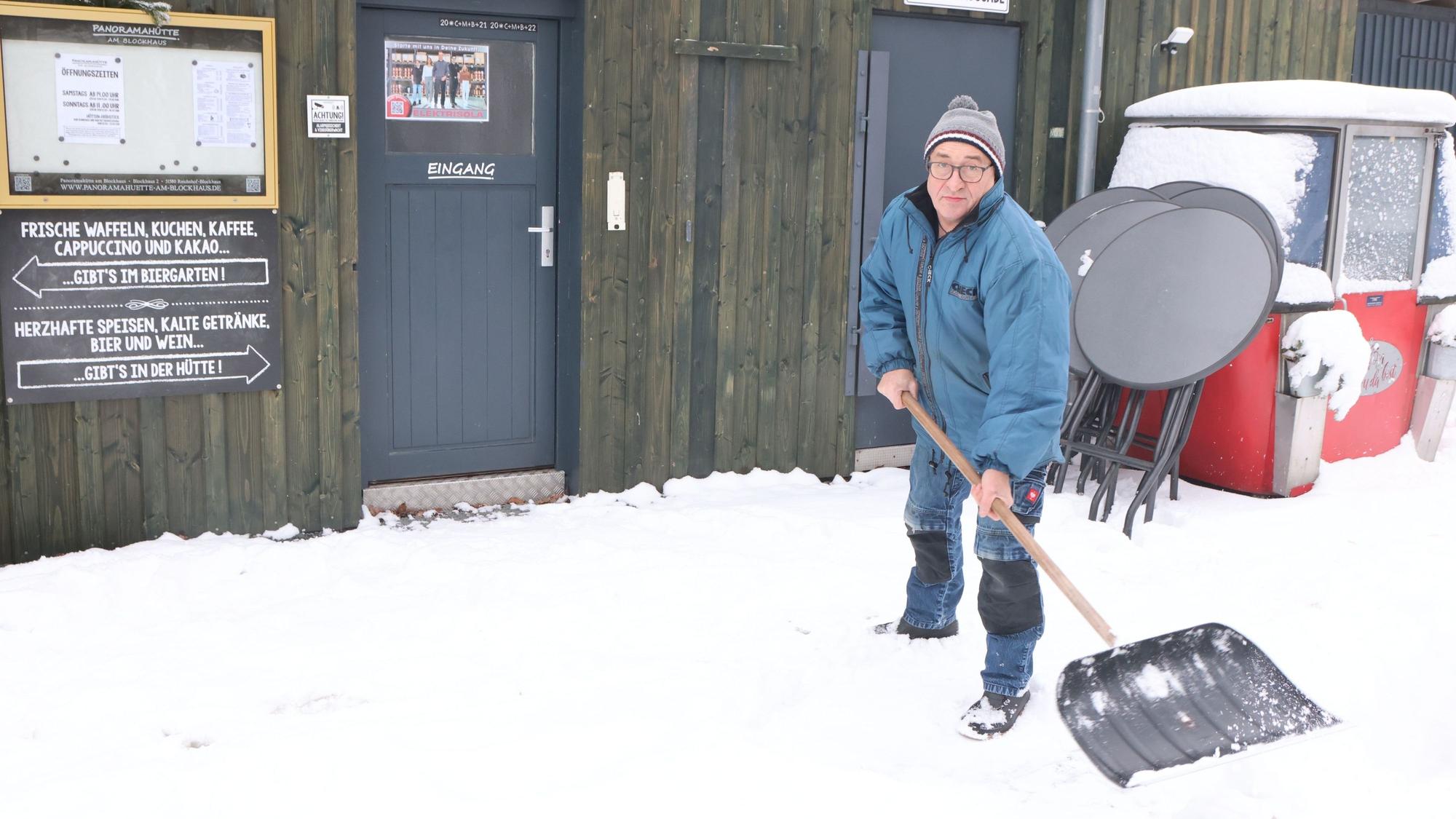 Ein Mann schaufelt vor einer Holzhütte Schnee.