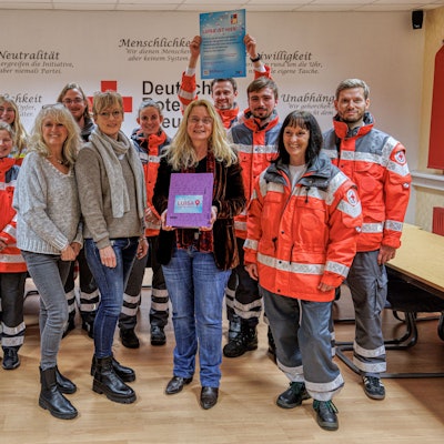 Das Team des DRK steht in neonroter Arbeitskleidung in einem Büro und hält Infomaterial in der Hand.
