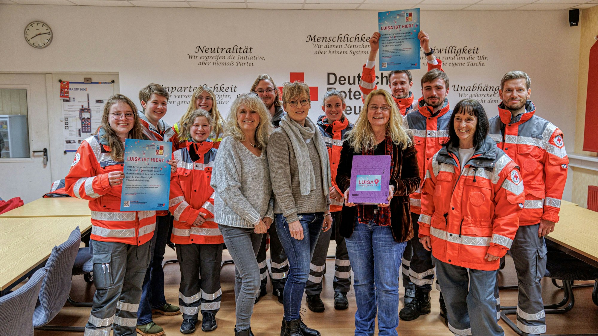 Das Team des DRK steht in neonroter Arbeitskleidung in einem Büro und hält Infomaterial in der Hand.