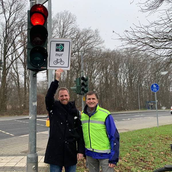 An einer roten Ampel stehen zwei Männer, einer hält ein Schild mit einem grünen Pfeil und dem Symbol eines Fahrrades in die Höhe.