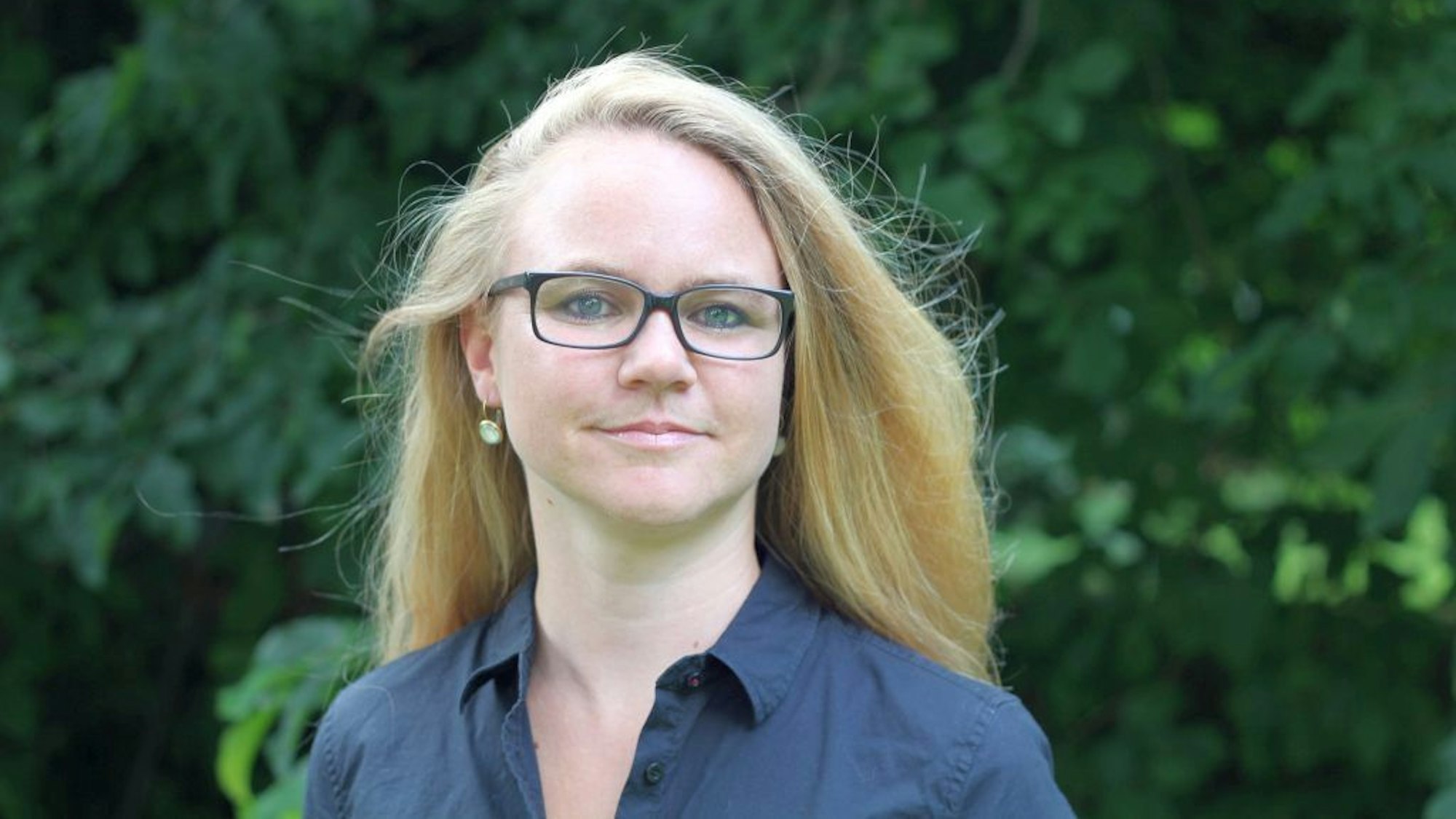 Sandra Vogel vor einem Hintergrund aus Bäumen. Sie hat lange blonde Haare und eine Brille.