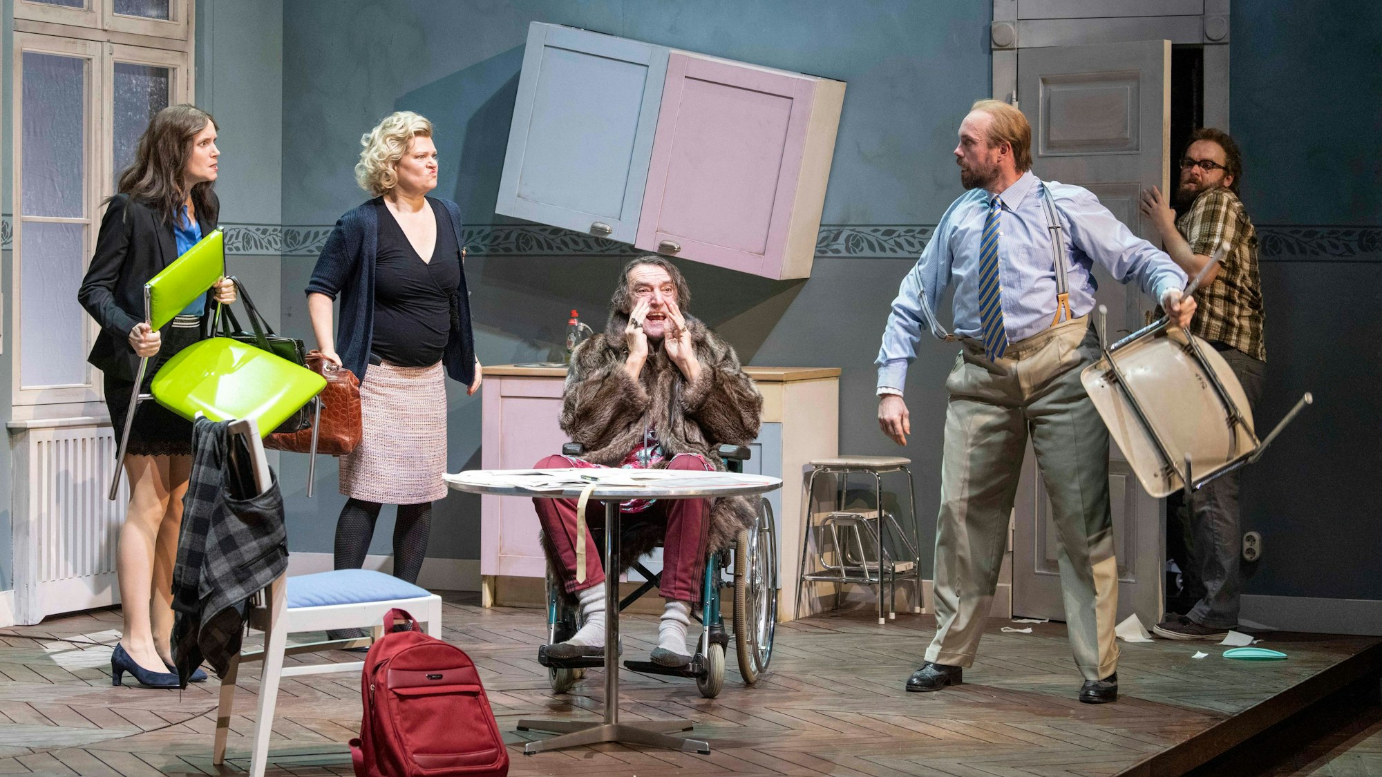 Premiere von „Der Haken“: Mehrere Personen auf der Bühne, die wie eine Küche eingerichtet ist. Im Hintergrund ein schräg hängender Hochschrank. Eine Frau und ein Mann mit Stühlen in der Hand.