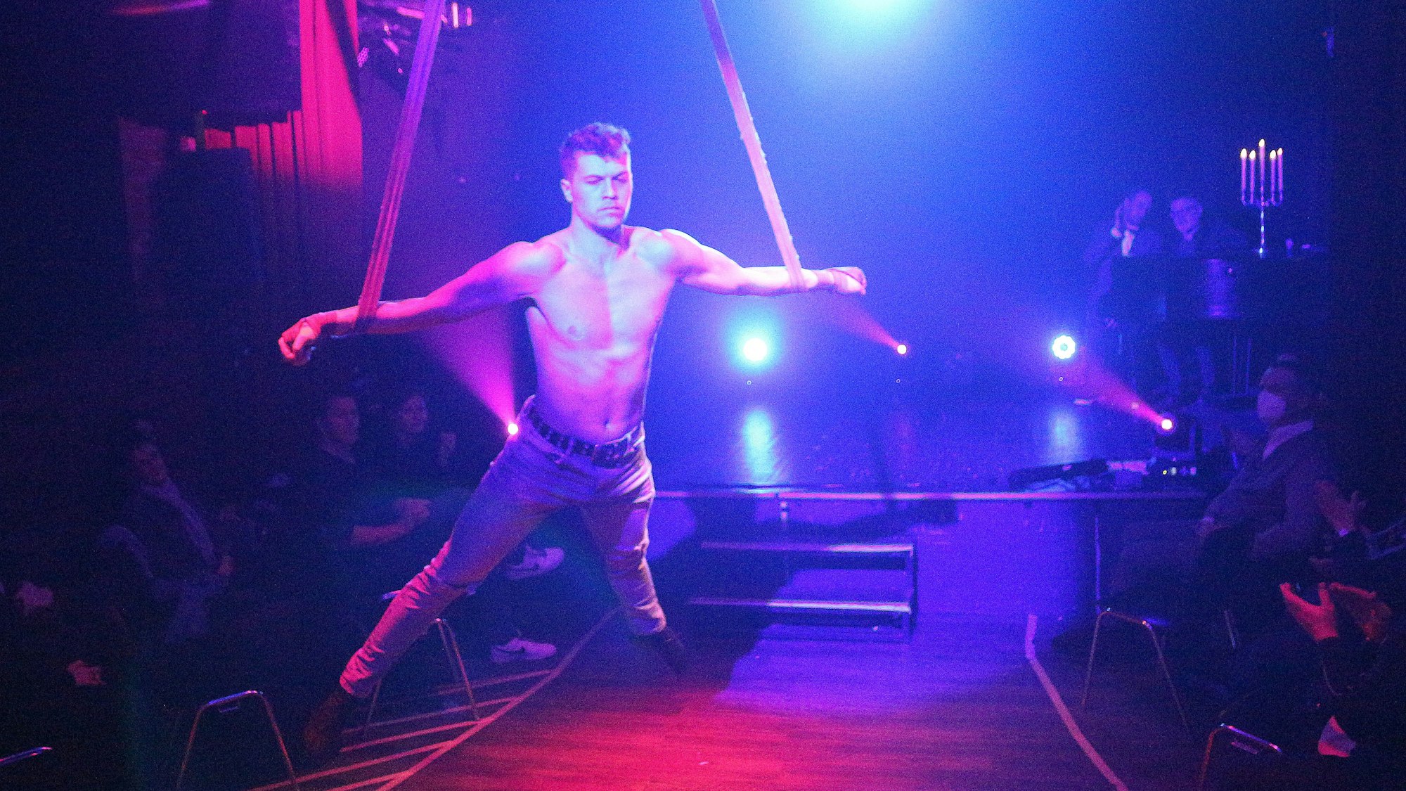 Ein Akrobat in Jeans und ohne Oberteil hängt mit ausgebreiteten Armen an Seilen in der Luft.