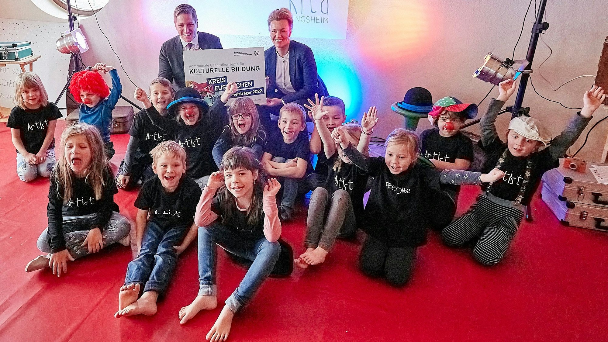 Eine Gruppe Kinder freut sich über das Preisgeld in Form eines großen Schecks, das eine Frau an einen Mann überreicht.