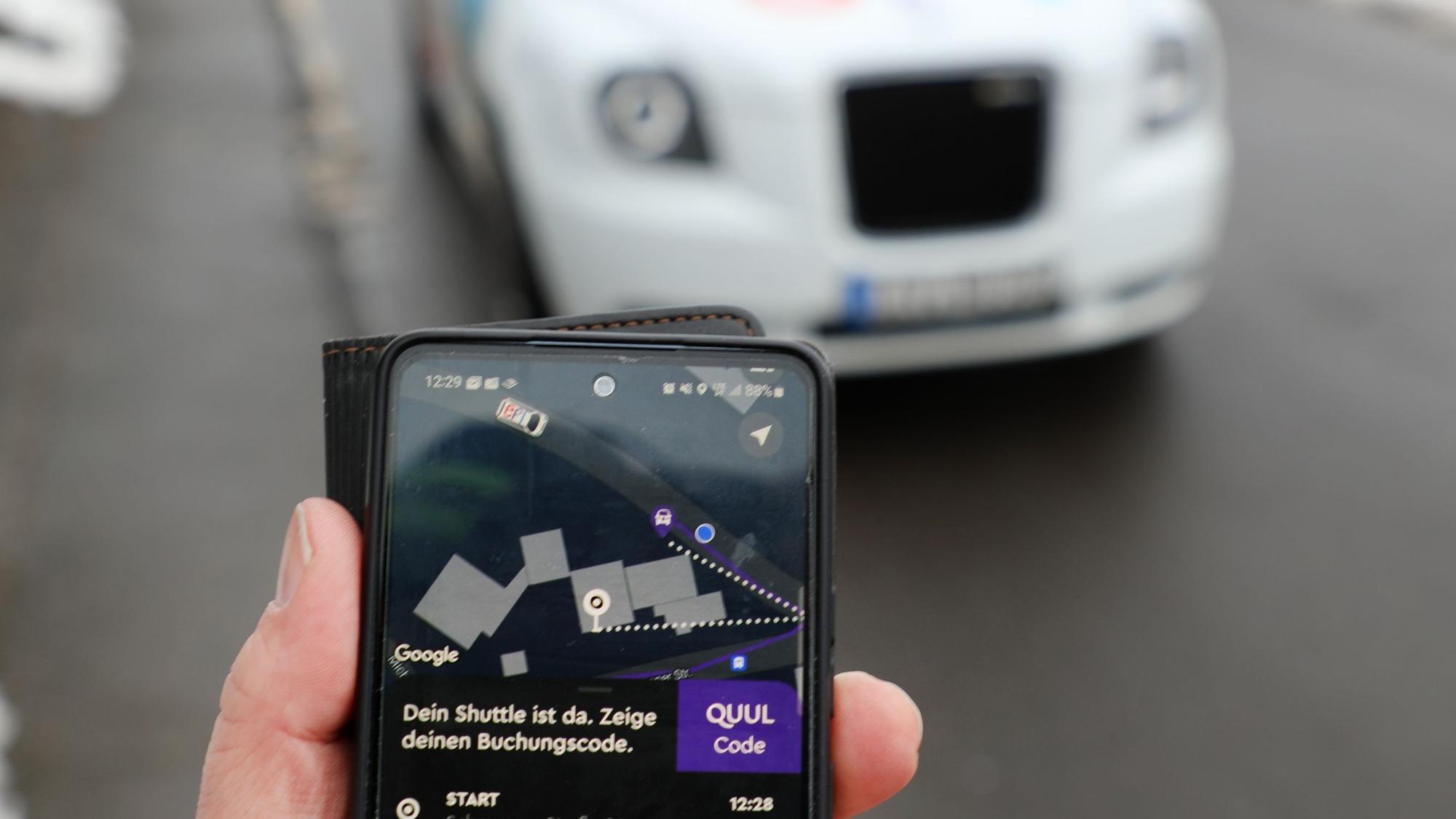 Eine Handy-App mit der Aufschrift „Dein Shuttle ist da“ ist vor einem gerade angekommenen „Efi“-Taxi zu sehen.