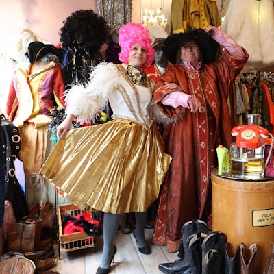 Eine Fau in einem gold-weißen Fransen-Dress und ein Mann in einer rot-goldenen Robe schunkeln in dem dem Second-Hand-Geschäft.