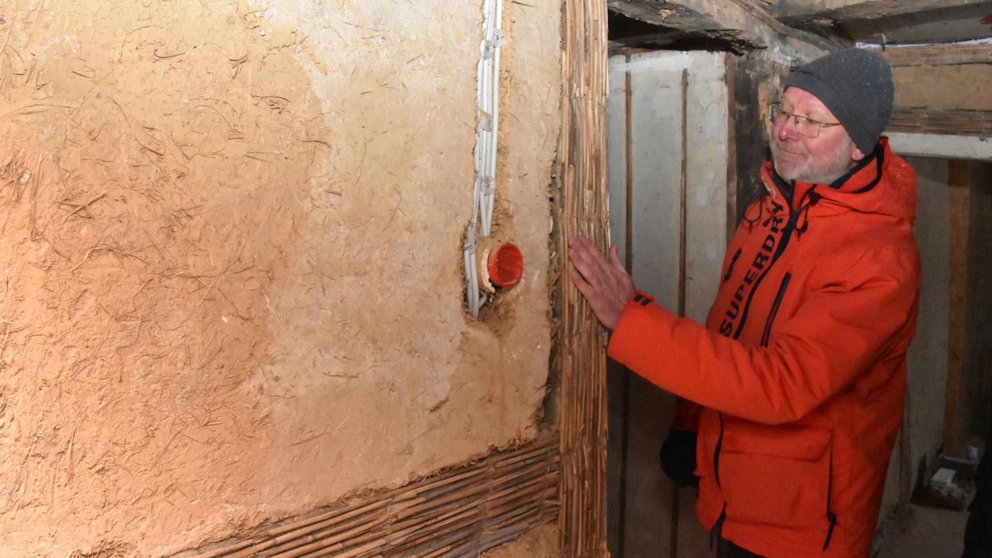 Ein Mann mit Anorak und Mütze steht an einer lehmverputzten Wand, an der an einer Stelle das Strohgeflecht sichtbar ist.