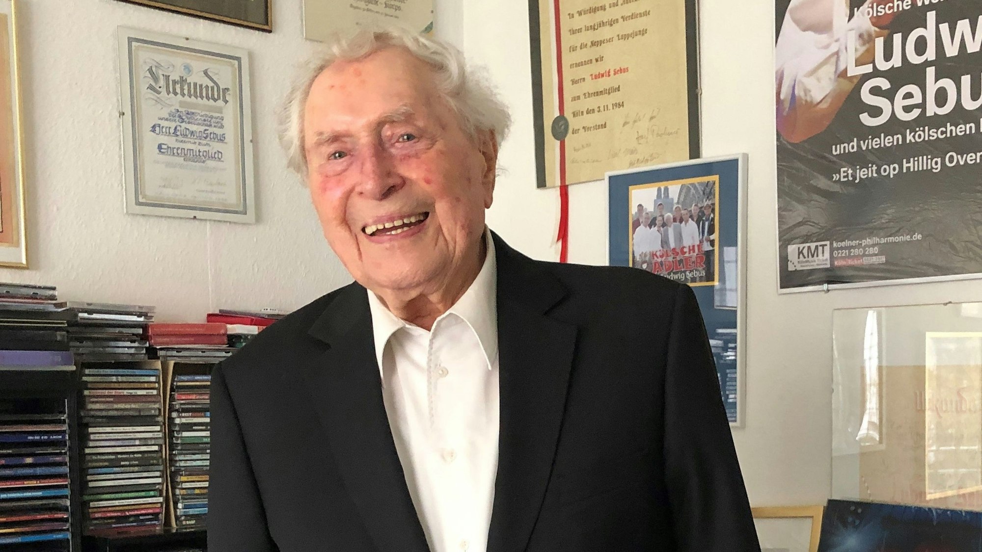 Bestens gelaunt empfing die „kölsche Legende“ Ludwig Sebus seine Gesprächspartnerinnen Daniela Janusch und Simone Fühles-Ubach in seinem Wohnzimmer. Sebus lacht und blick in die Kamera der Gesprächspartnerinnen, die nicht im Bild zu sehen sind. Wohl aber jede Menge CDs in Regalen und Urkunden an den Wänden der Wohnzimmerecke.